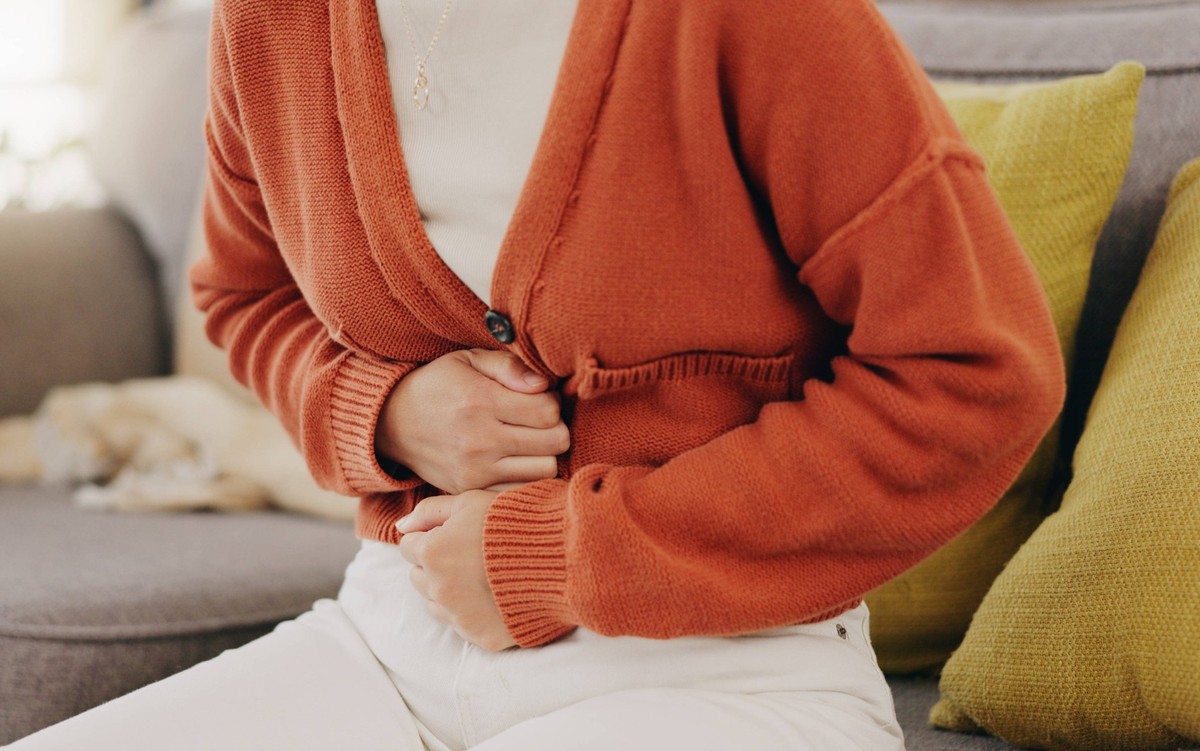 Stomach ache, cramps and hands of woman with abdomen pain due to constipation, menstruation or ibs issue. Sick, home and person suffering and holding belly in a house lounge, couch and living room. PU ...