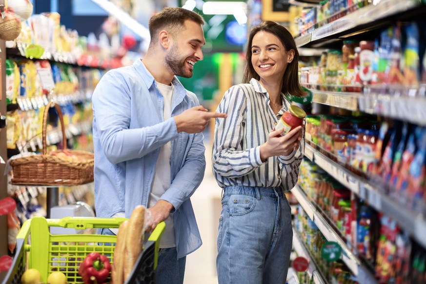 Grocery Shopping. Young Smiling Family Couple Buying Groceries In Supermarket Store Indoors. Two Buyers Standing With Shopping Trolley Cart In Aisles Choosing Food Products And Laughing