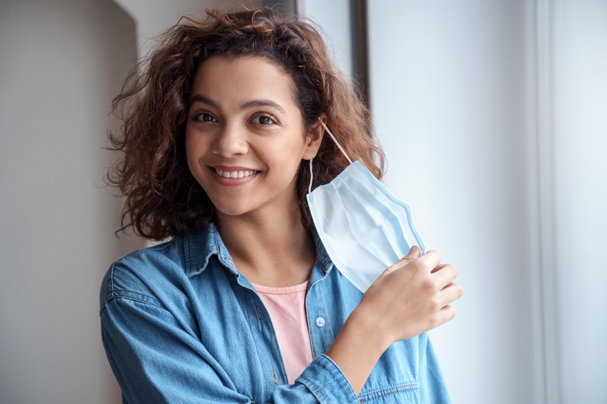 Happy Hispanic young woman takes off protective mask and looking at camera indoors while corona virus pandemic. Quarantine, and social distance concept.