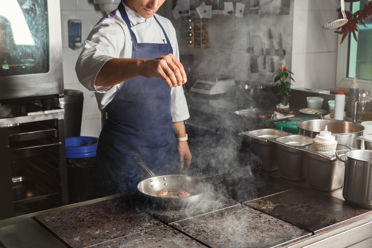 Male chef roasting and seasoning meat at professional restaurant kitchen. Man in uniform busy at work