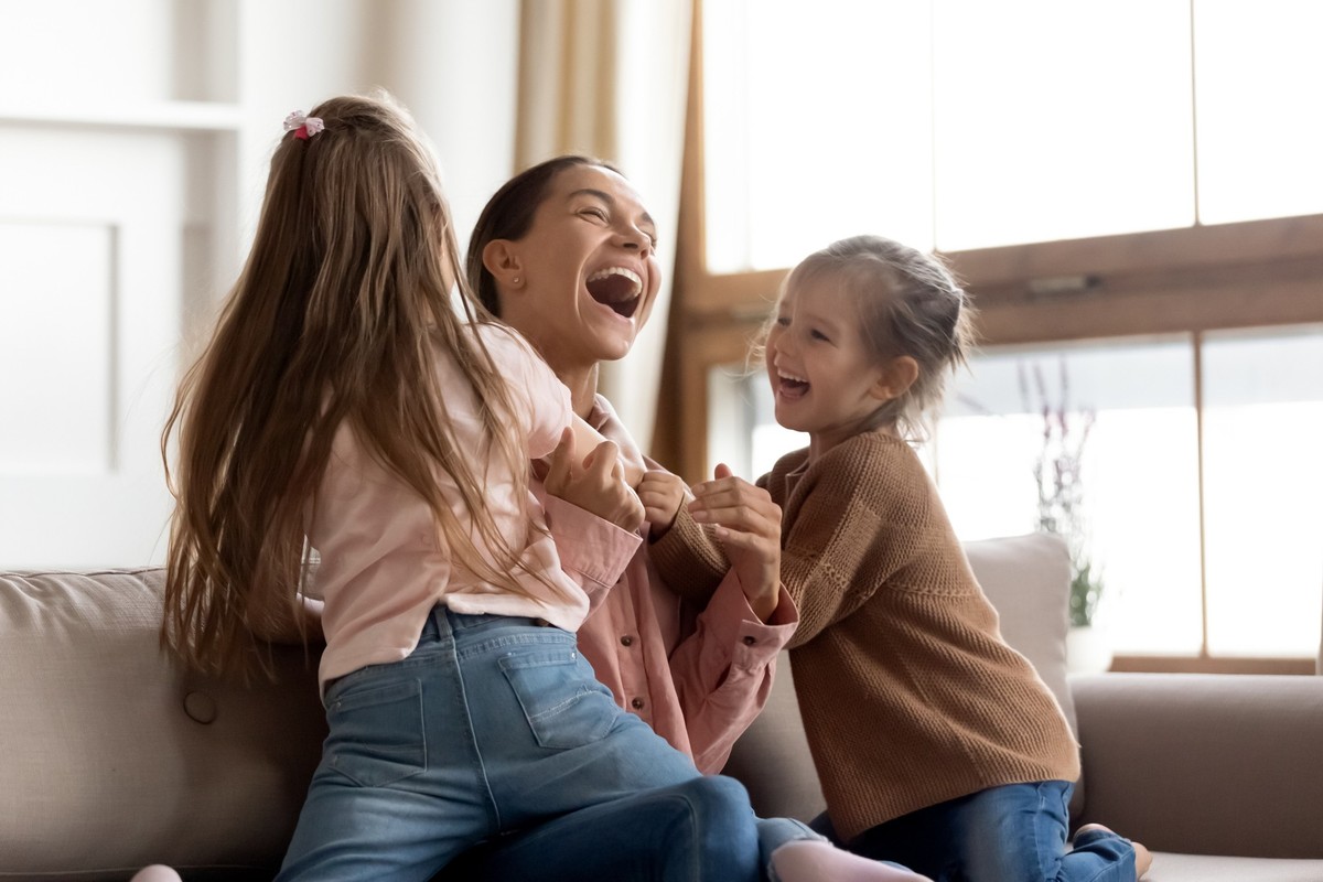 Cheerful positive family cute small funny kids children daughters enjoying tickling playing lifestyle game with young happy mum embracing laughing having fun relaxing sitting on sofa at home together