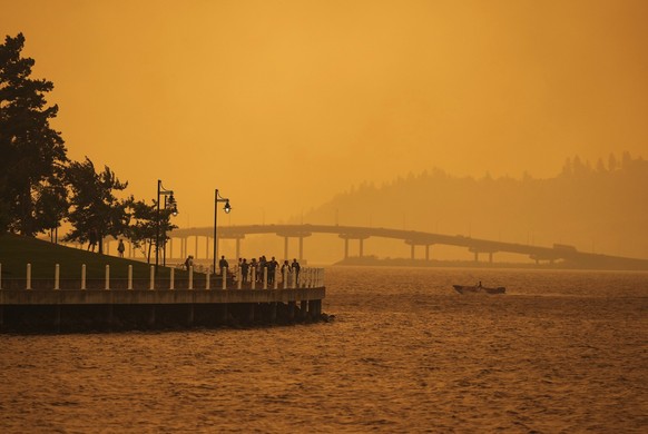 18.08.2023, Kanada, Kelowna: Eine Person fährt in einem Boot an Menschen vorbei, die auf der Promenade spazieren gehen, während der Rauch des McDougall Creek Waldbrandes das Gebiet am Okanagan Lake in ...