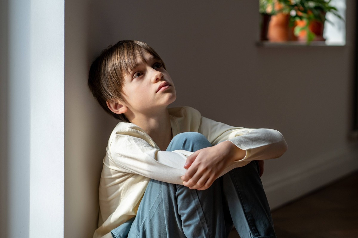 Einsamer Junge schaut traurig auf und umarmt seine Knie, während er allein zu Hause am Fenster sitzt. Kind psychologisches Trauma Lonely little boy sadly looking up hugging knees sits near window at h ...