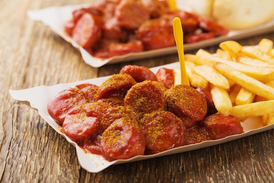 Traditional German currywurst, served with chips on disposable paper tray. Wooden table as background.