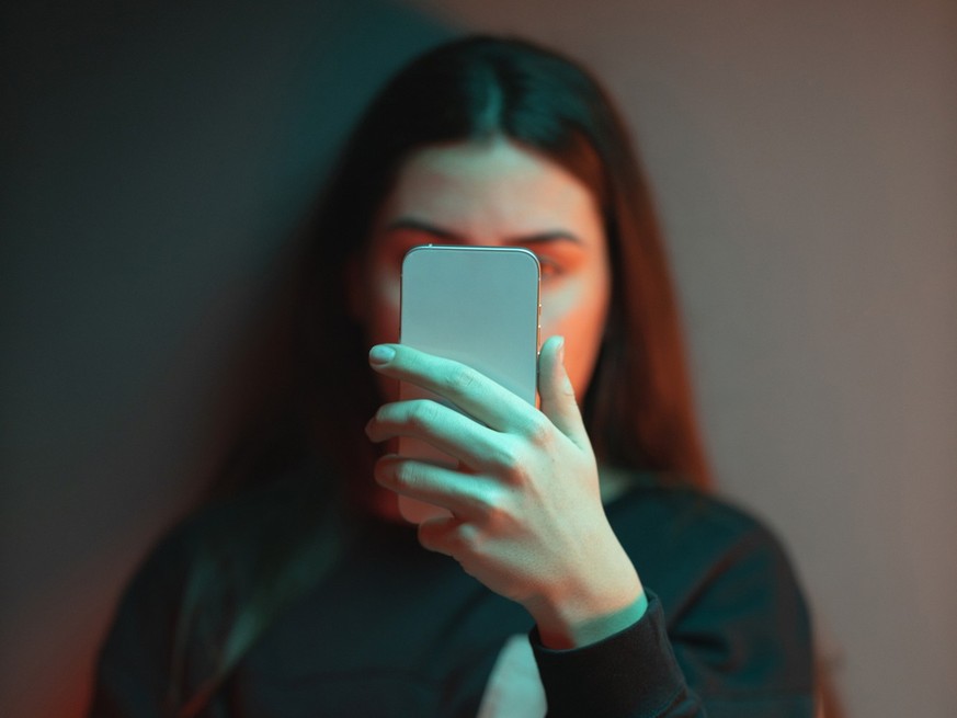 Close-up of a depressed teenage girl sitting on the bed in her room lightened with orange and teal neon lights and scrolling social media.