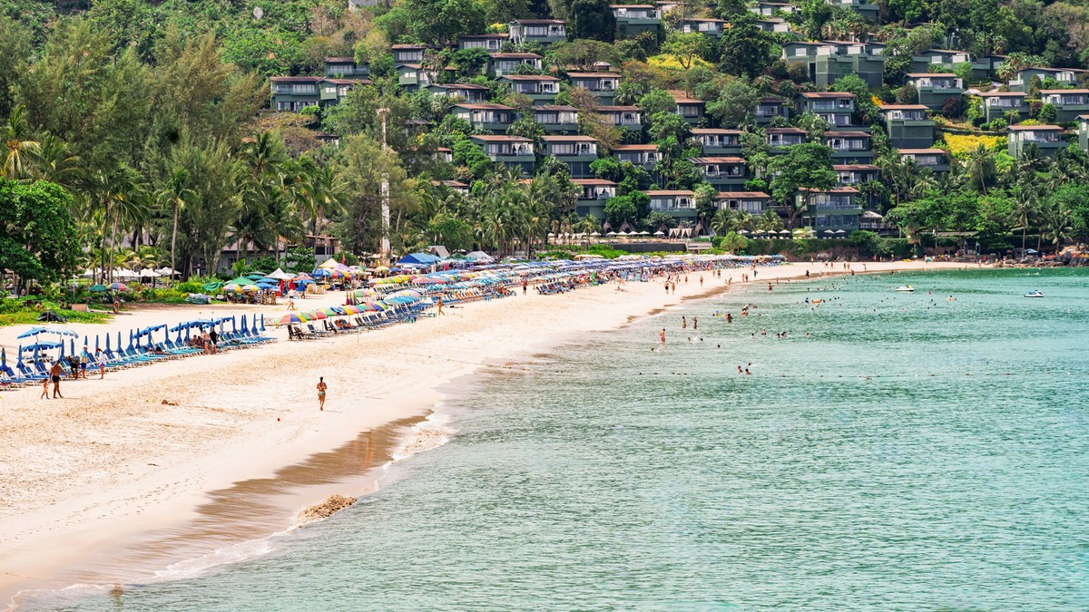 A scenic beach in Phuket, Thailand, lined with umbrellas and sunbeds, bustling with people enjoying the sun, and hillside villas overlooking the turquoise sea and lush greenery Model Released IrinaChe ...