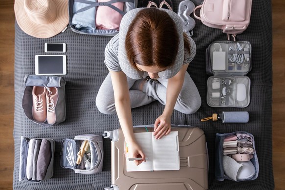 Smiling tourist woman packing suitcase to vacation writing paper list sitting on bed at home. Female getting ready to travel trip organization things storage in comfy cases bags use konmari method