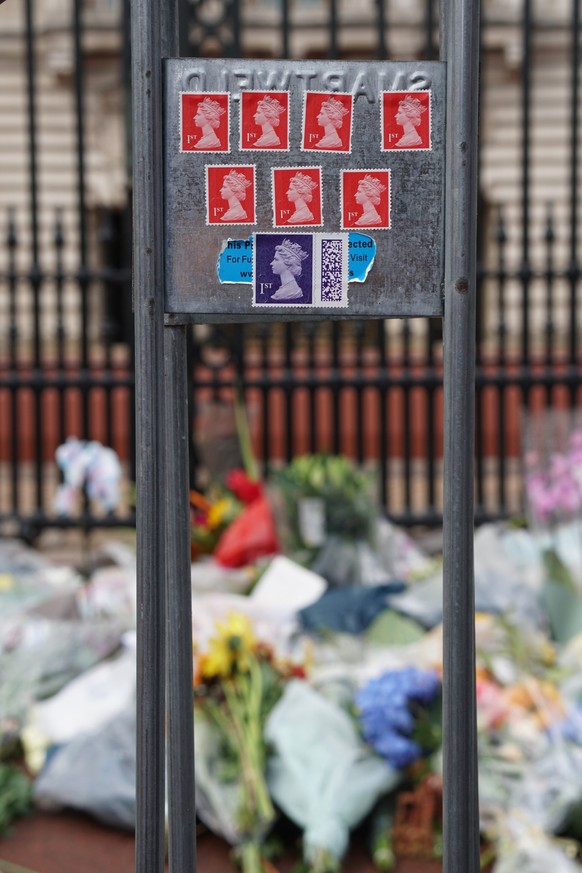 09.09.2022, Großbritannien, London: Briefmarken von Königin Elizabeth II. kleben an einem Zaun am Buckingham-Palast - im Hintergund sind zahlreiche Blumensträuße zu sehen. Die britische Königin Elizab ...