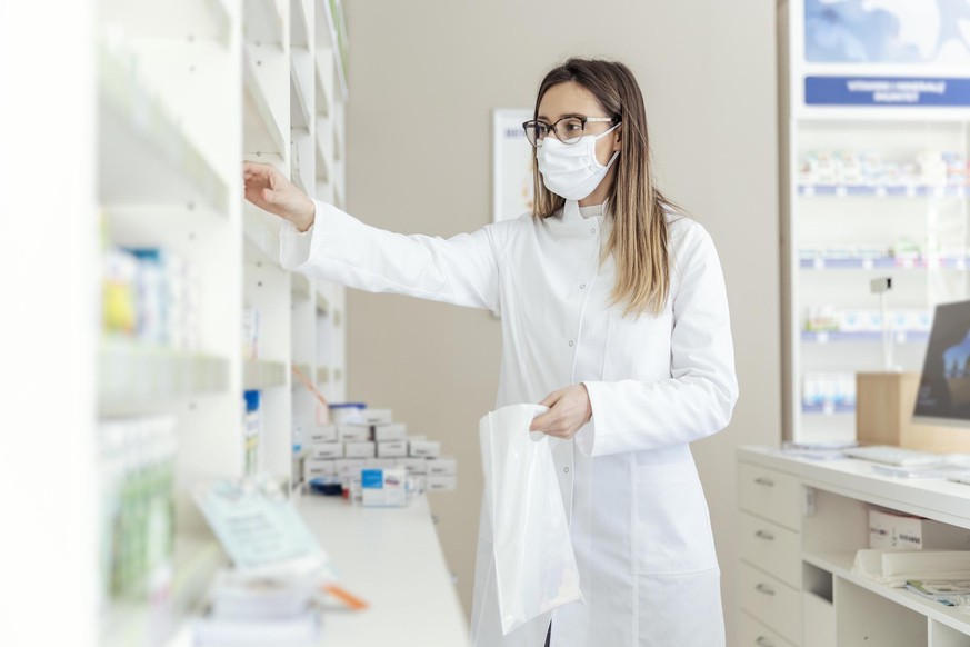 Work in a pharmacy. Packing medicines in a bag. Female employed as a pharmacist and dressed in a white uniform with a protective mask on her face in bags packing boxes of medicines and supplements