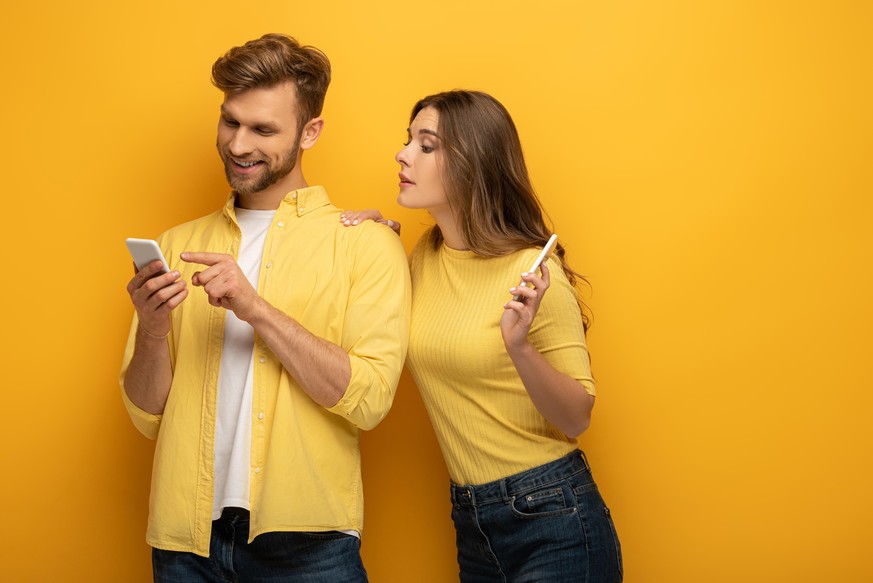 Smiling man pointing with finger at smartphone near girlfriend on yellow background