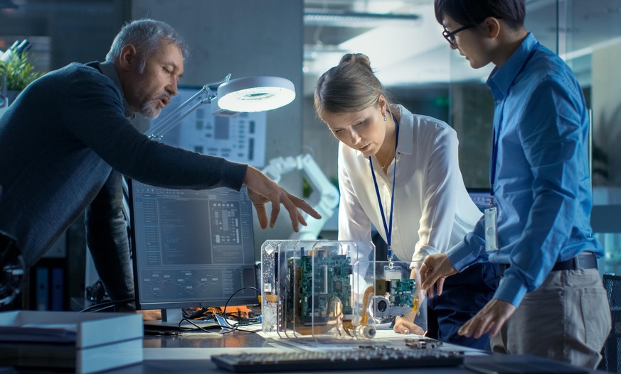 Team of Computer Engineers Lean on the Desk and Choose Printed Circuit Boards to Work with, Computer Shows Programming in Progress. In The Background Technologically Advanced Scientific Research Cente ...