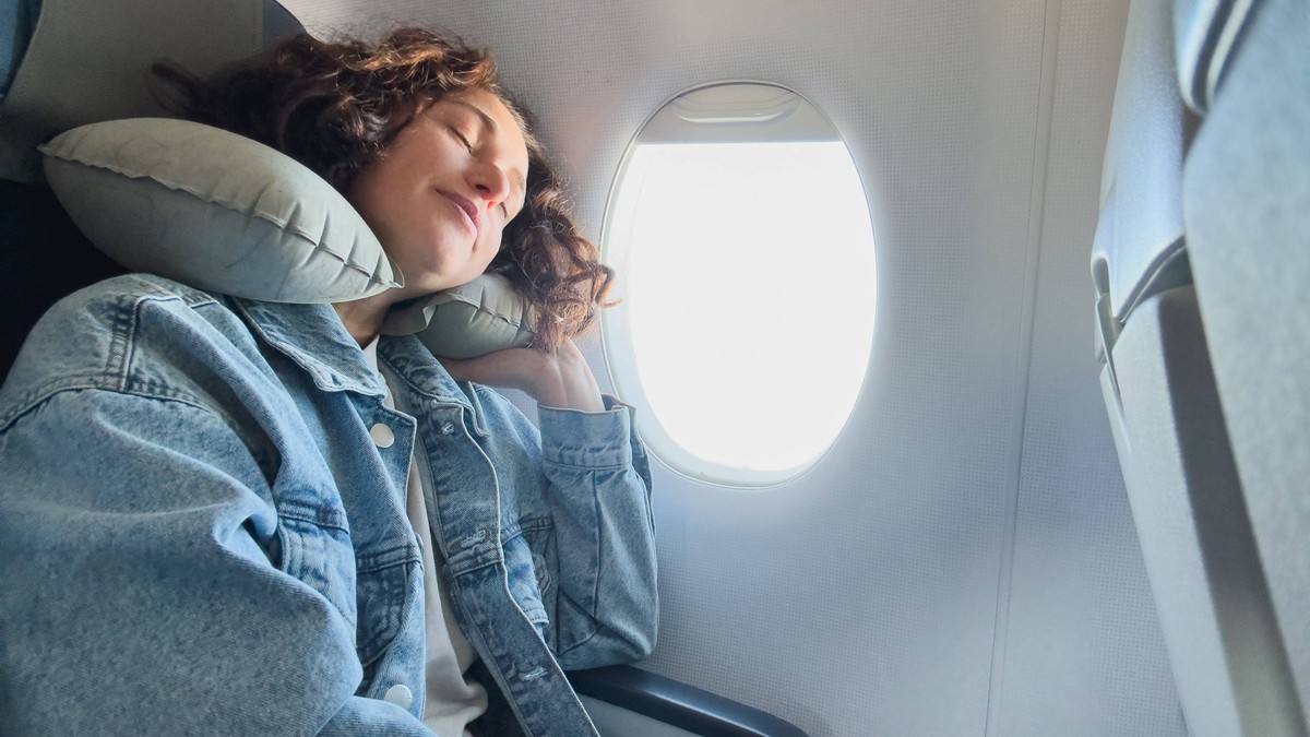 Rest on plane. Young woman napping sitting by porthole in business jet using neck pillow for travel. Passenger girl flying in plane. Girl sleeping in airplane going on trip vacation. Traveling girl. R ...