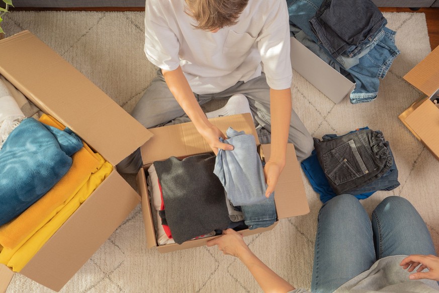 Woman and child sorting clothes and packing into cardboard box. Donations for charity, help low income families, declutter home, sell online, moving moving into new home, recycling, sustainable living ...