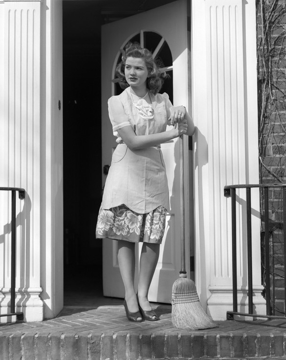 UNITED STATES - Circa 1940s: Woman Wearing Apron With Broom In Open Front Door Leaning On Broom Looking Unhappy Sad Sweep Clean Housewife.