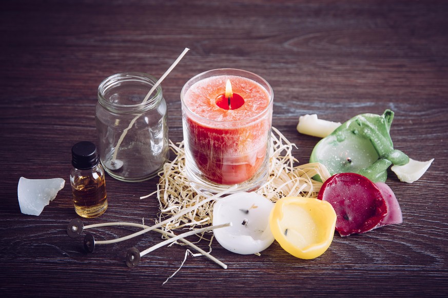 Reusing old candles leftovers and making melting a new one: various ingredients on table: candle wicks, glass jar, old candles wax, aroma oil. New candle in glass in center.