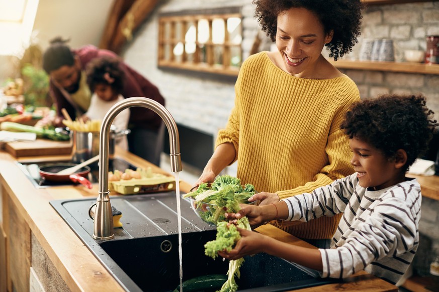 Happy African American boy assisting his mother and washing lettuce while preparing food in the kitchen. Focus is on mother.