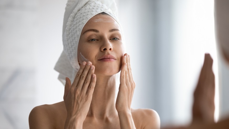 Closeup head shot pleasant beautiful woman applying moisturizing creme on face after shower. Smiling young pretty lady wrapped in towel smoothing perfecting skin, daily morning routine concept.