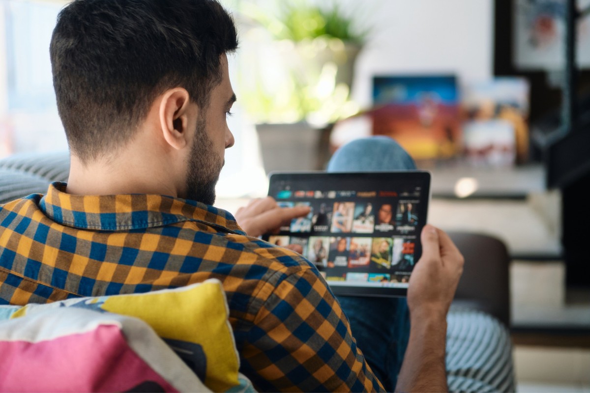 Young white man lying down on sofa in living room at home searching for on line films or TV series to watch on his tablet.