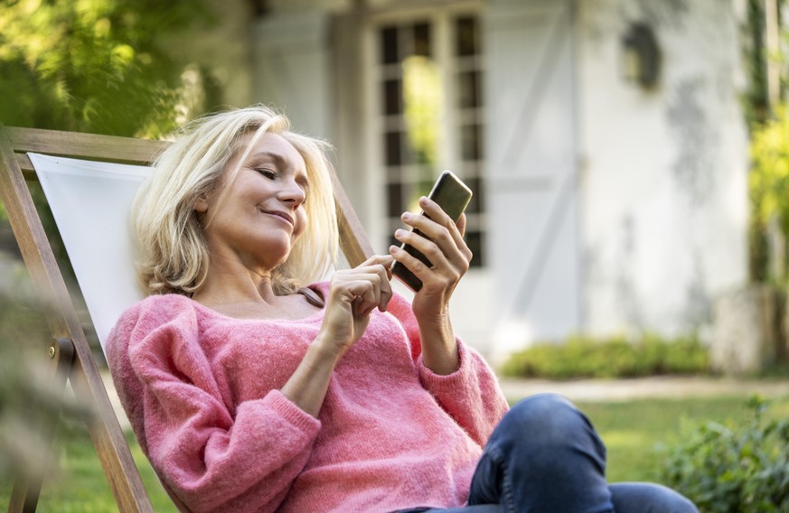 Smiling mature woman using smartphone while sitting on deckchair || Modellfreigabe vorhanden