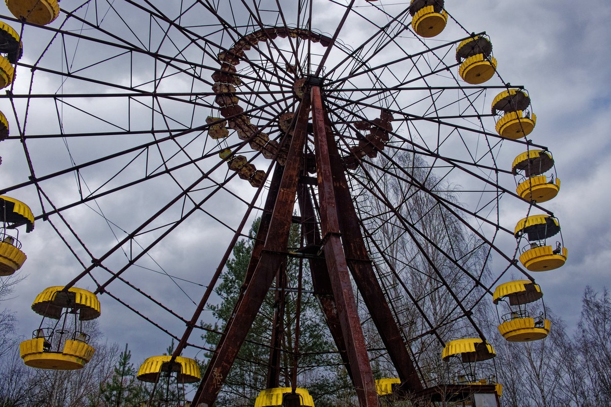 A rusted Ferris wheel with yellow cabins against a cloudy sky. xkwx ferris wheel rust cabins sky trees clouds gondolas forest metal structure yellow coloration weathering abandonment amusement park ou ...