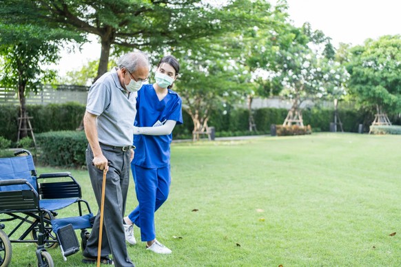 Asian young woman nurse at nursing home take care and support disabled senior elderly man on walker or cane at backyard. Caregiver doctor and old patient wearing mask to prevent from Covid-19 pandemic