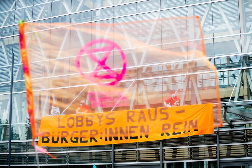 Umweltaktivisten sitzen neben einem Banner bei einer Blockade der Landesvertretung von Nordrhein-Westfalen (NRW) auf einem Vordach, wobei sie durch eine transparente Fahne der Extinction-Rebellion-Bew ...