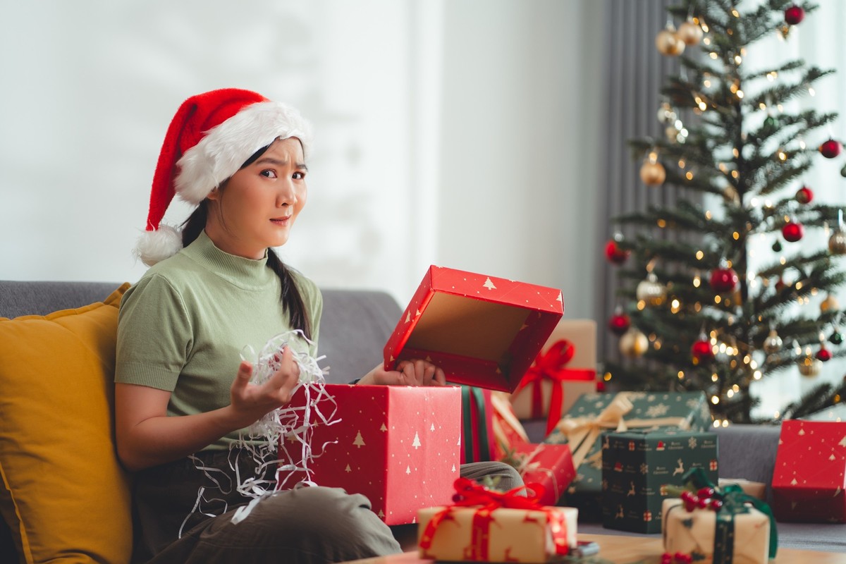 Asian woman sitting in living room decorated with Christmas tree feel disappointed when she sees the gift in the box. Looking at camera.