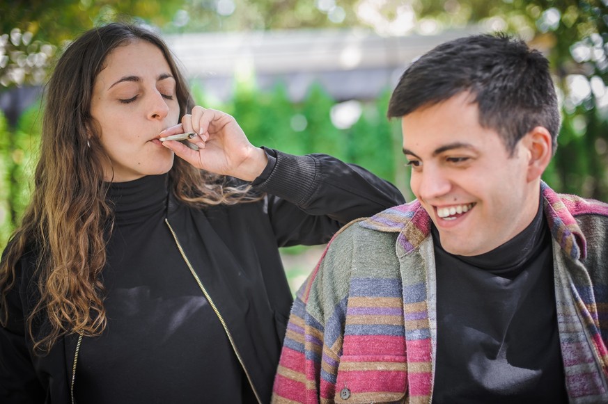 Young relaxed and smiling happy young couple, woman and man, smoking cannabis marijuana ganja or hashish joint in public park outdoor. Female and male smoke thc green grass together. Real life