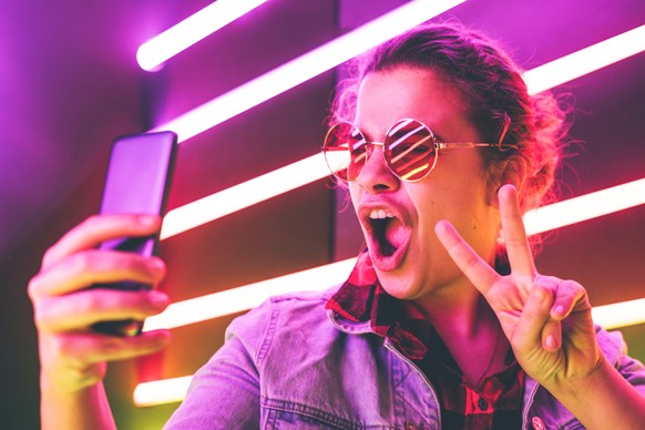 Portrait of modern young girl in round glasses making selfie at the neon light. Shows sign pis.