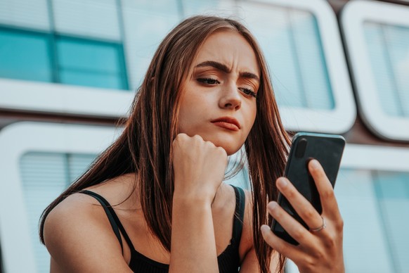 Reading Bad News on Mobile Phone. Young teenage woman checking her messages and social media post on her mobile phone. Looking worried and questioning reading the bad news and messages on her smart ph ...