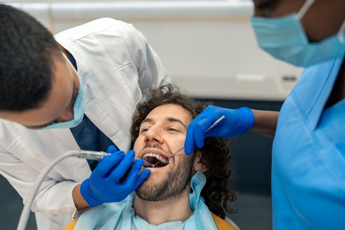 Young man having dental drill procedure at dentist&#039;s office. Female dentist specialist and dental assistant working on patient&#039;s teeth using dental drill, removing dental plaque.