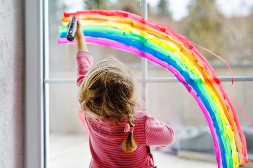 Adoralbe little toddler girl with rainbow painted with colorful window color during pandemic coronavirus quarantine. Child painting rainbows around the world with the words Let&#039;s all be well
