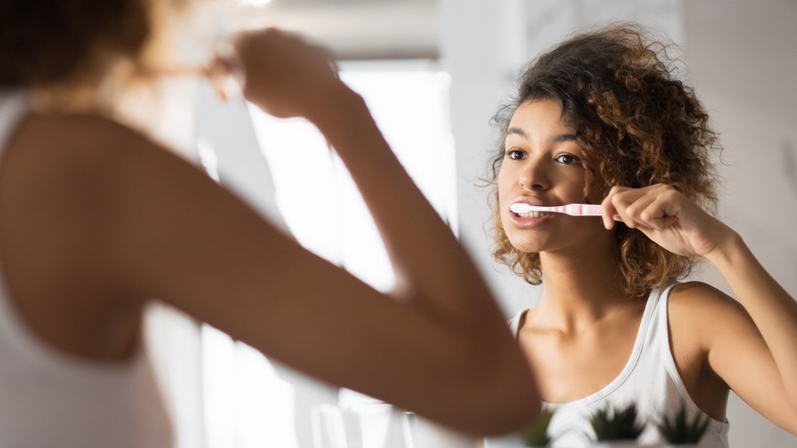 African American Girl Brushing Teeth With Toothbrush Looking In Mirror In Bathroom. Dental Care Concept. Panorama, Selective Focus