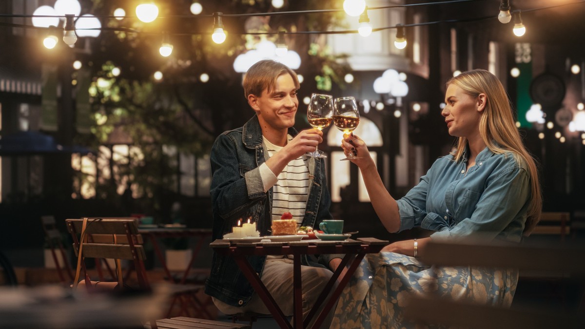 Young Beautiful Couple is Having a Romantic Date at a Cafe at Night. Happy Man and Woman Sitting on a Terrace, Raising Wine Glasses, Toasting while Having Lovely Conversations