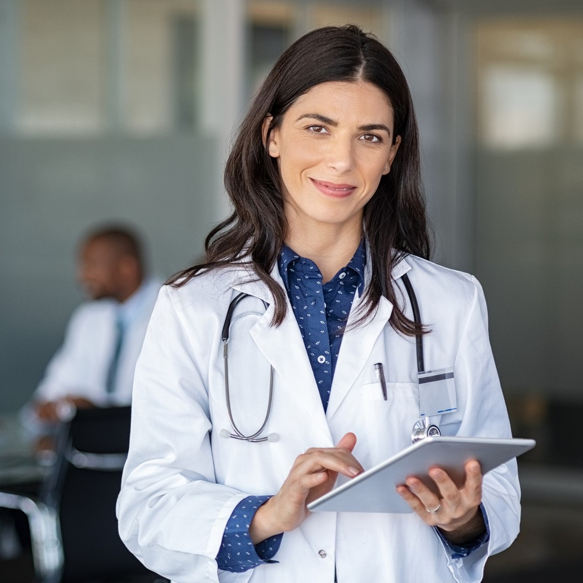 Portrait of beautiful mature woman doctor holding digital tablet and looking at camera. Confident female doctor using digital tablet with colleague talking in background at hospital. Latin nurse in la ...