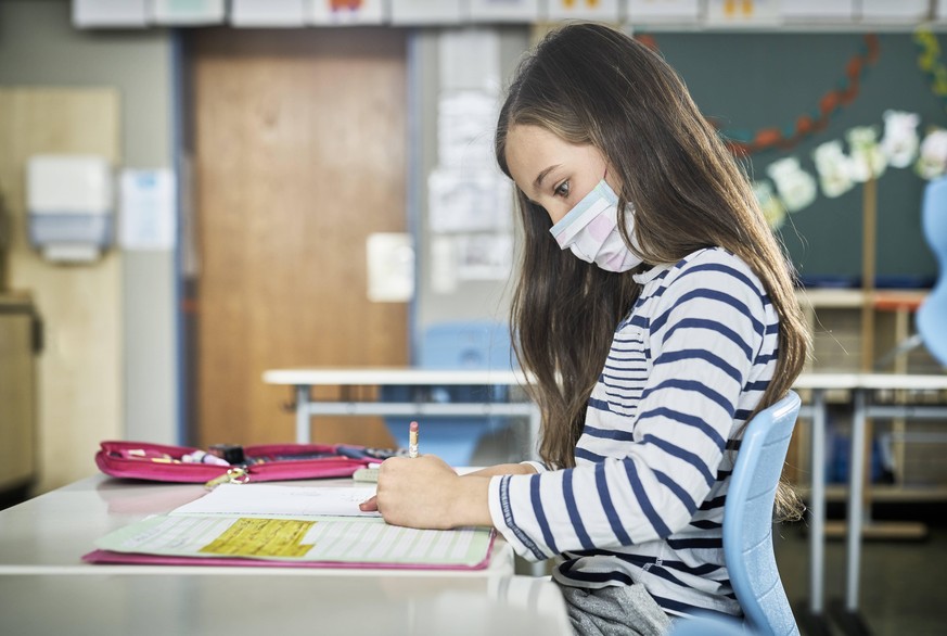 Girl wearing mask in classroom writing in workbook model released Symbolfoto property released DIKF00516