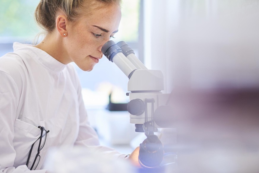 a female research scientist is analysing a sample on her microscope in a microbiology lab . the lab is brightly lit with natural light . Blurred glassware at side of frame provides copy space .