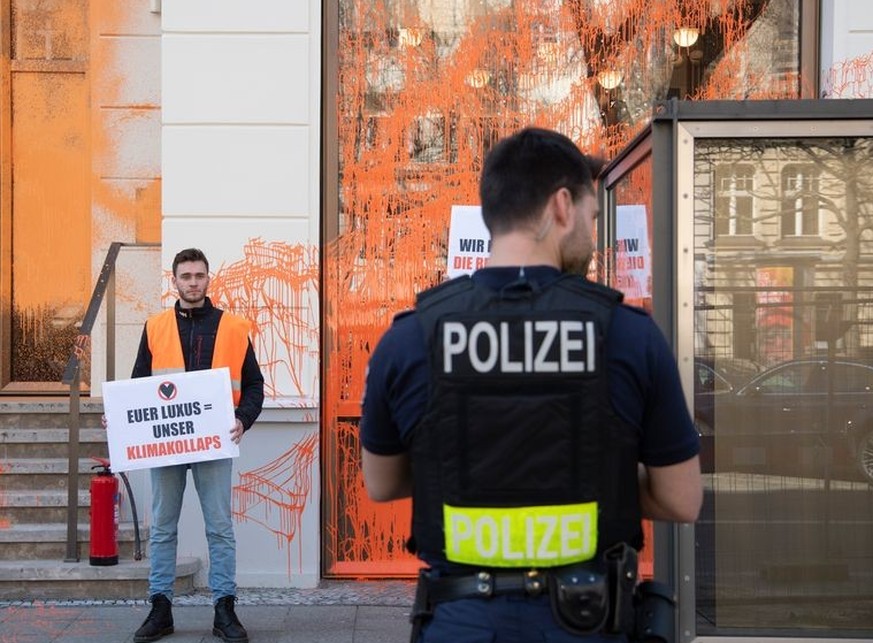 22.04.2023, Berlin: Ein Aktivist der Umweltschutzgruppe Letzte Generation hält vor einem Luxusgeschäft ein Plakat. Ein Polizist beobachtet ihn. Zuvor hatte der Aktivist die Fassade mit Farbe besprüht. ...