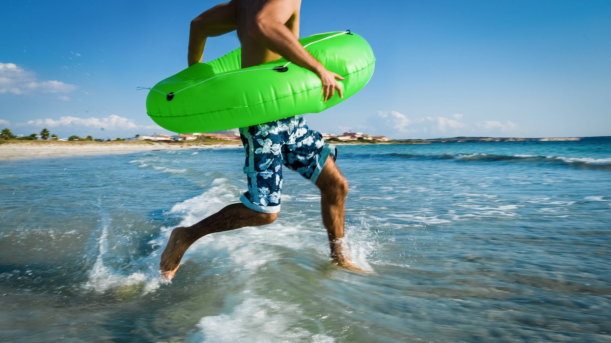 Man wearing swimming shorts and carrying green inflatable ring running into the Mediterranean Sea. Putzu Idu Italy Copyright: xx