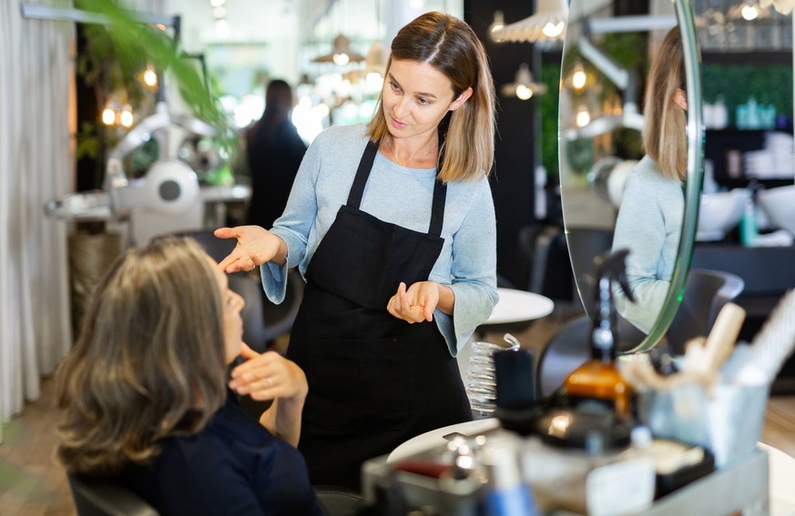 Portrait of young woman professional hair stylist talking to elderly female client in salon, choosing new hairdo