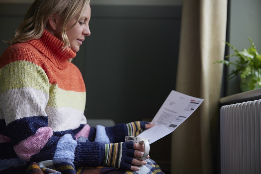 Woman In Gloves With Hot Drink And Bill Trying To Keep Warm By Radiator During Cost Of Living Energy Crisis