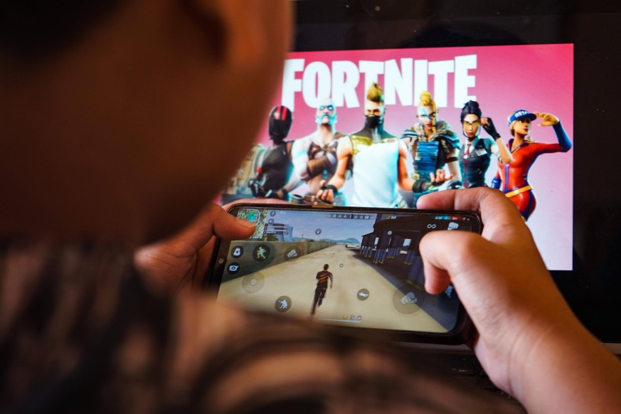 August 15, 2020, Makassar, South Sulawesi, Indonesia: A child is playing a game on a cellphone with a picture of the Fortnite game on the computer screen in the background. Apple and Google announced  ...