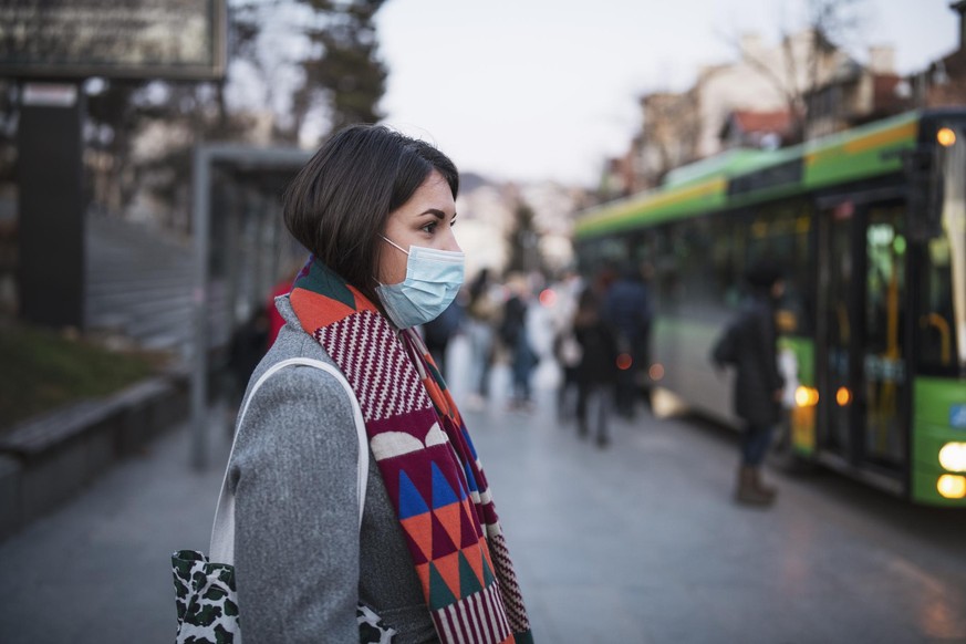 Side view of young woman with protective face mask in town, she standing in front at the bus stop and waiting for her transport after work.