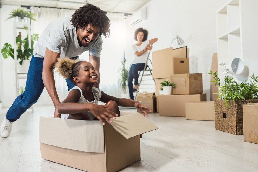 Little preschooler daughter sitting in cardboard box, father rolling her. Smiling mother looking them.