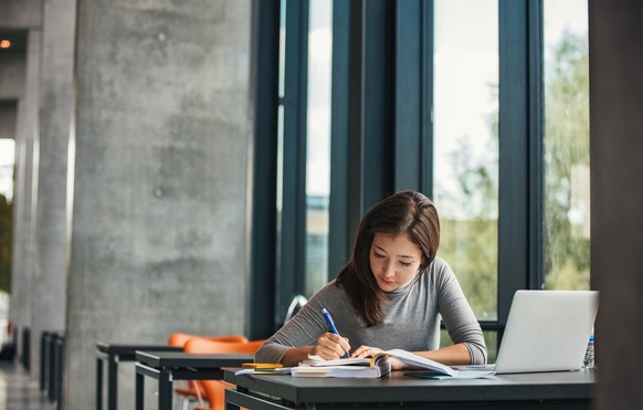 Shot of young asian female student sitting at table and writing on notebook. Young female student studying in library.