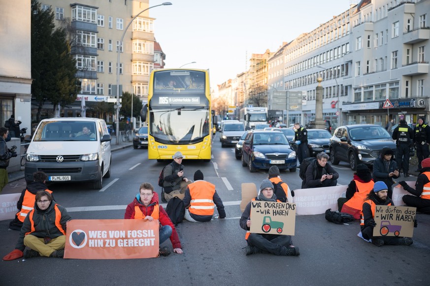 10.01.2024, Berlin: Aktivisten der Klimaschutzgruppe Letzte Generation sitzen bei einer Straßenblockade mit Schildern mit aufgemalten Traktoren auf der Straße. Aktivisten nehmen damit Bezug auf die Ak ...