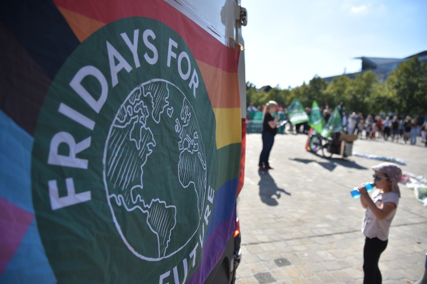 15.09.2023, Magdeburg: Eine Regenbogenfahne mit Logo von Fridays for Future hängt an der Rückseite eines kleinen Transportwagens. Im Hintergrund ist eine Rednerin zu sehen, die beim sogenannten «Klima ...