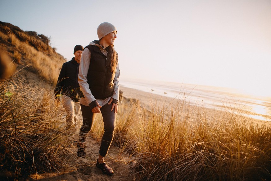 A senior couple explores a beach in Oregon state, enjoying the beauty of sunset on the Pacific Northwest coast. They hike up a sand dune, the ocean visible in the background.