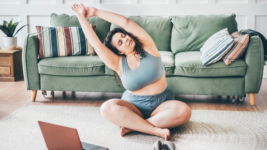 Curly haired overweight young woman in top and shorts turns on online yoga training and practices exercises on floor mat against green sofa by wall