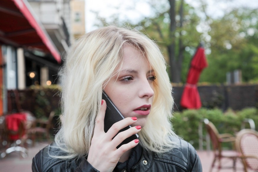 Junge Frau mit langen blonden Haaren telefoniert mit ernstem Gesichtsausdruck mit dem Handy in der Öffentlichkeit. 27.04.2017 || Modellfreigabe vorhanden